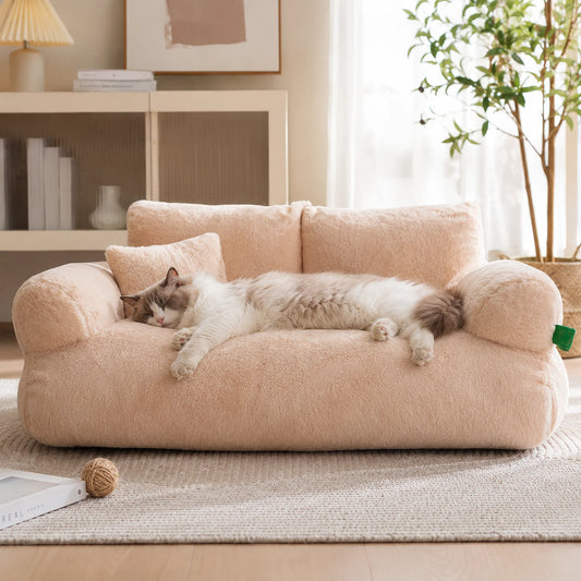 A fluffy cat lies stretched out on a plush, pale blush-pink pet sofa in a bright, modern living room with a rug, shelving, and a potted plant in the background.